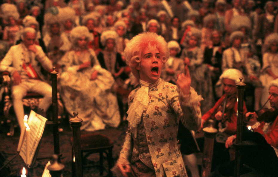 cena z filmu Amadeusz – młody Wolfgang Amadeusz Mozart w peruce i stroju z epoki prowadzi orkiestrę podczas występu w XVIII-wiecznym pałacu, otoczony publicznością w bogatych kostiumach i perukach; w tle muzycy i arystokracja, wszystko oświetlone ciepłym światłem świec.